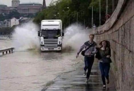 Imagem de um caminhão prestes a passar por uma poça d'água e molhar os pedestres que estão na via lateral.