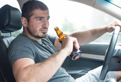 é melhor soprar ou se recusar a fazer o teste do bafômetro? Imagem de um homem dentro do carro segurando uma garrafa de bebida alcoólica na mão direita e a outra apoiada no volante, olhando fixamente para frente.